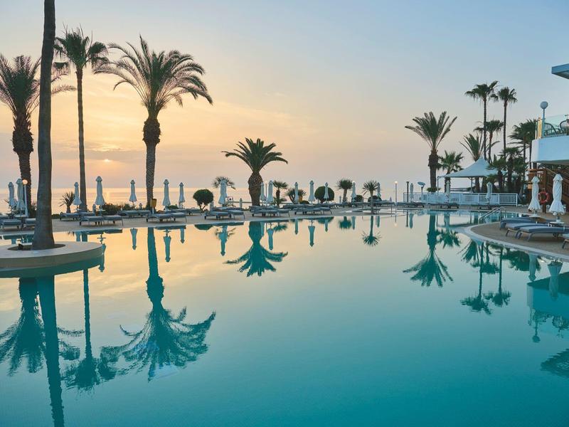 Großer Hotelpool mit Palmen und Sonnenuntergang am Meer im Hintergrund