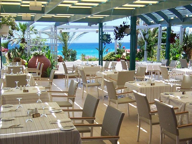 Offenes Restaurant mit weißen Tischen, Stühlen und Blick auf das blaue Meer bei Sonnenlicht.