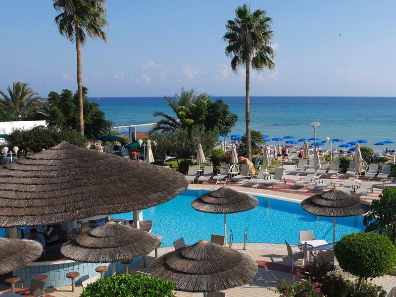 Schwimmbad mit Sonnenschirmen und Liegestühlen, dahinter Palmen und blauer Meereshorizont.