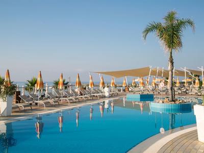 Großer Pool mit Liegestühlen und Sonnenschirmen neben dem Meer und Palmen.