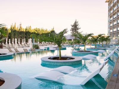 Pool mit Liegestühlen und Palmen neben einem Hotelgebäude bei Sonnenuntergang.