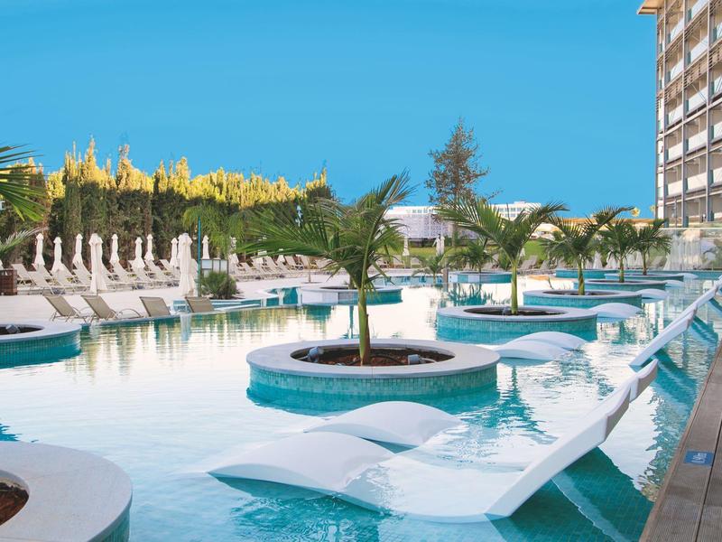 Außenpool mit Palmen und Liegestühlen neben einem Hotelgebäude unter klarem Himmel.