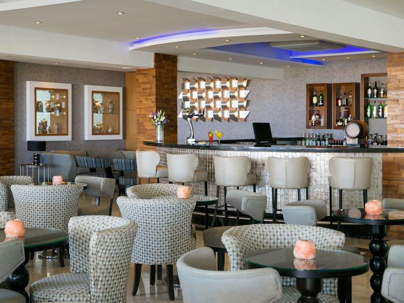 Moderne Hotelbar mit vielen gepolsterten Sesseln und einem beleuchteten Deckenakzent.