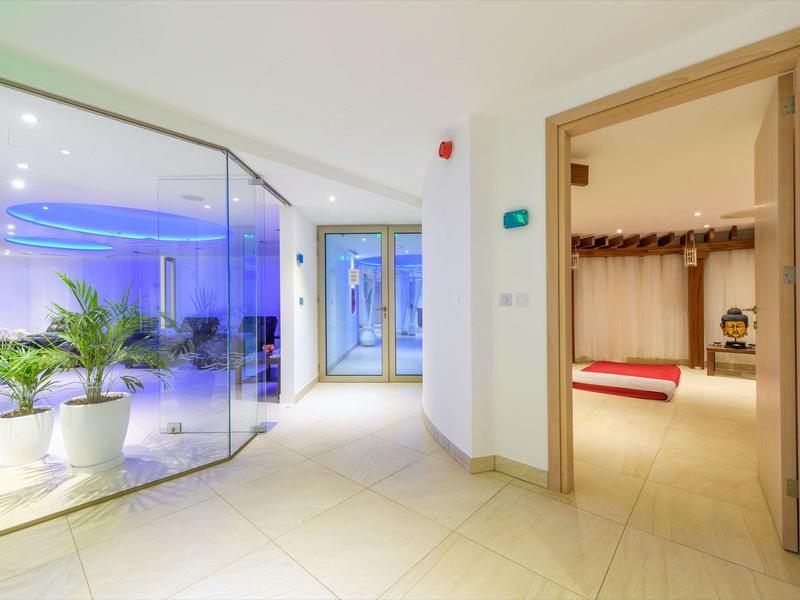 Heller Hotelflur mit Glastrennwand und Blick in einen gemütlichen Raum mit rotem Kissen.