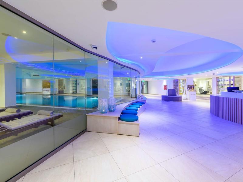 Moderner Wellnessbereich mit blauer Beleuchtung, Lounge-Sesseln und Schwimmbad mit Glasabtrennung.