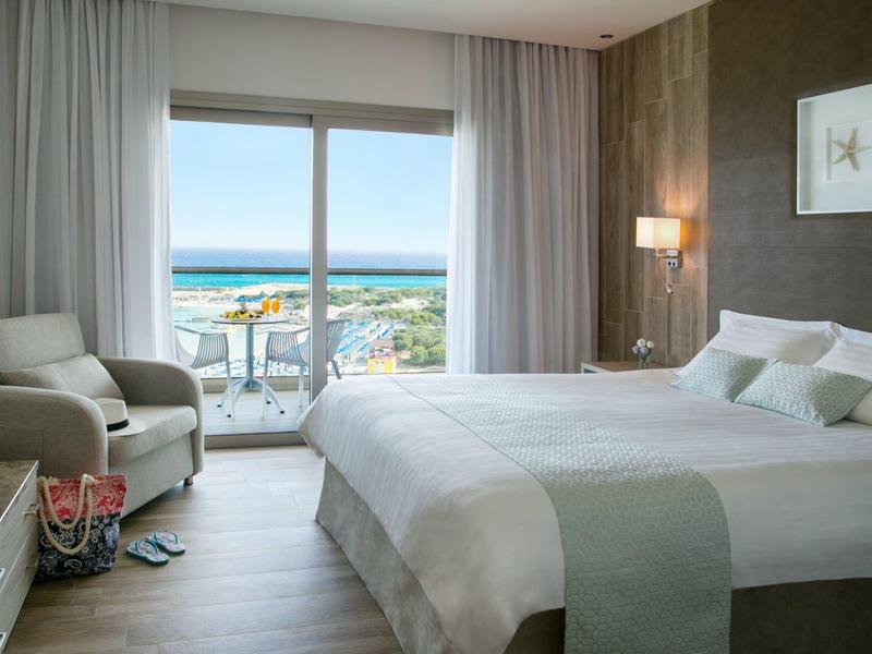 Hell eingerichtetes Hotelzimmer mit Doppelbett und Blick auf das Meer durch bodentiefe Fenster.