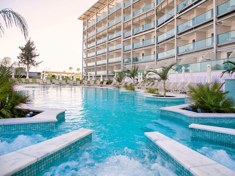 Großer Pool mit Palmen neben einem modernen Hotelgebäude bei Sonnenuntergang.