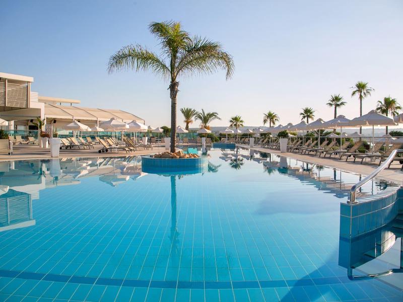 Großer Hotelpool mit Palmen, Liegestühlen und klarem blauem Wasser an sonnigem Tag.