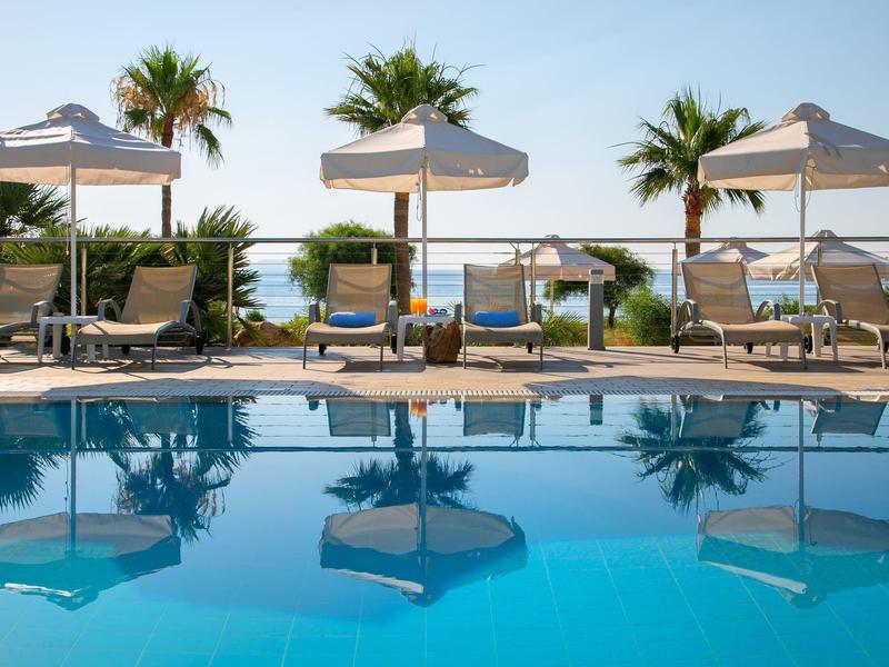 Pool mit Sonnenliegen und Sonnenschirmen, Blick auf Palmen und Meer im Hintergrund.