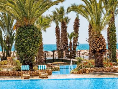 Vista di una piscina con sdraio, palme e un ponte verso il mare.