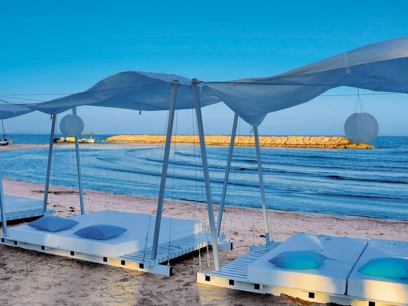 Strand mit modernen, weißen Holzplattformen, blauen Kissen und blauem Sonnenschutzdach bei klarem Himmel.