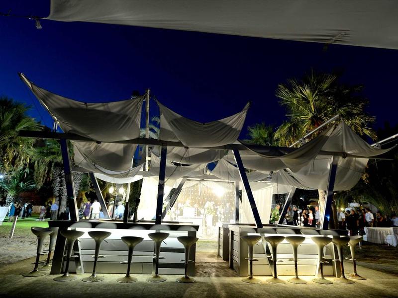 Beleuchtetes Zelt mit weißen Segeln und Stühlen in tropischer Nachtumgebung.