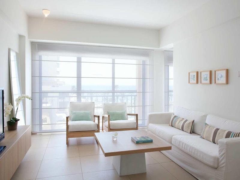 Helles Wohnzimmer mit großem Fenster, weißen Wänden, zwei Sesseln, Sofa und Holztisch.