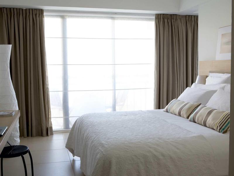 Schlichtes Schlafzimmer mit großem Bett, weißen Bettlaken, braunen Vorhängen und schlichtem Nachtisch.