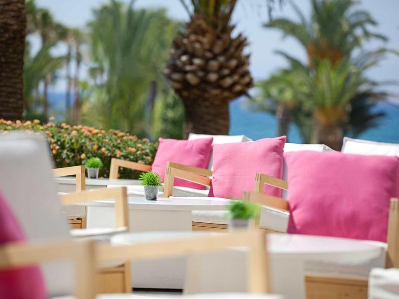 Outdoor-Sitzbereich mit weißen Möbeln, rosa Kissen und Palmen im Hintergrund am Meerufer.