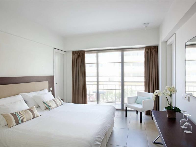 Helles Schlafzimmer mit großem Bett, weißen Wänden, Sitzstuhl und breitem Fenster mit braunen Vorhängen.