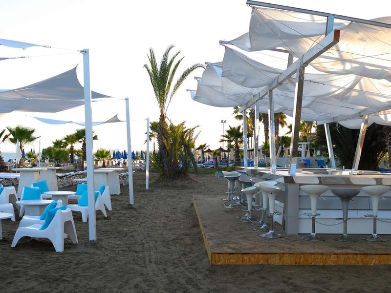 Strandbar mit weißen Sitzmöbeln und blauen Kissen unter weißen Sonnensegeln am Abend.