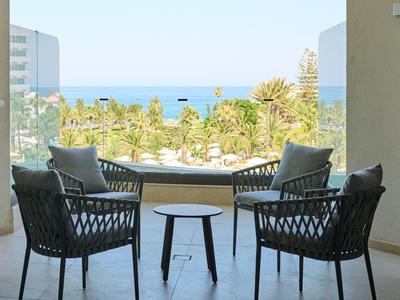 Balcone con tavolo e quattro sedie vista su palme e mare in una giornata limpida.