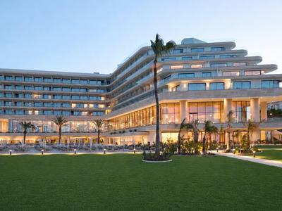 Grande hotel moderno con ampie vetrate e balconi illuminati al crepuscolo.