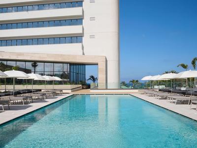 Terrazza moderna con piscina, lettini e ombrelloni bianchi accanto a un edificio alberghiero bianco.
