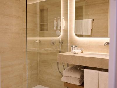 Bagno moderno in hotel con doccia a filo, specchio illuminato e asciugamani appesi.