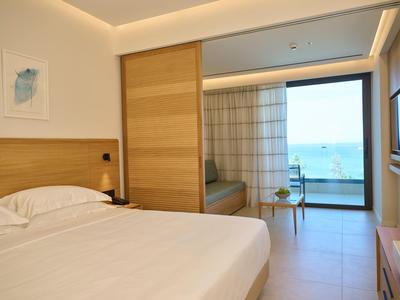 Camera d'hotel luminosa con letto matrimoniale, divano e vista mare attraverso finestre in vetro.