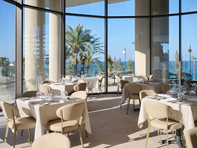 Ristorante luminoso con grandi finestre, vista su palme e mare, tavoli apparecchiati.