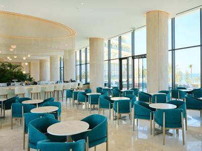Area lounge d'hotel luminosa e moderna con sedie blu, tavoli rotondi e grandi finestre.