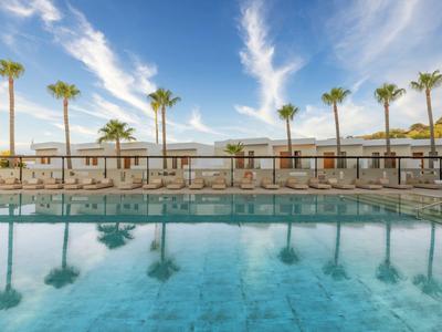 Vista de una piscina tranquila con palmeras y un edificio de hotel al fondo bajo un cielo azul.