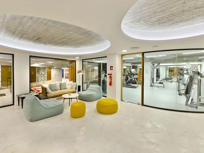 Moderner Lounge-Bereich mit hellen Sofas, runden gelben Hockern und großem Fitnessraum hinter Glaswänden.