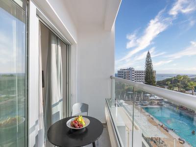 Balkon mit Glastrennwand, schwarzem Tisch mit Obstschale, Blick auf Pool, Bäume und Gebäude am Meer.