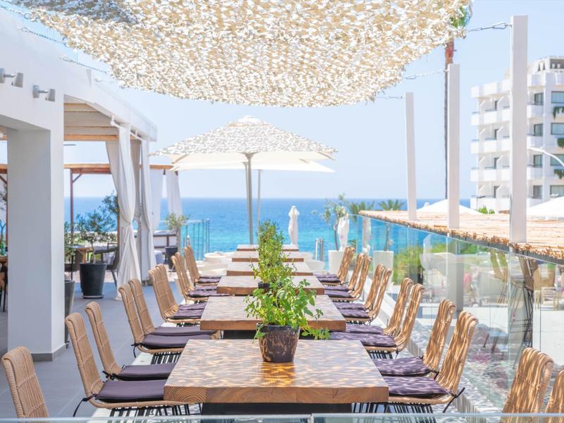 Terrassenrestaurant mit Holztischen, Pflanzen, Sonnenschirmen und Meerblick an einem sonnigen Tag.
