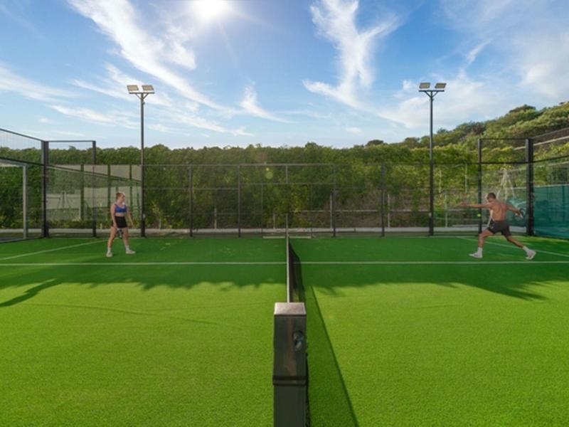 Zwei Personen spielen Tennis auf einem grünen Platz im Freien bei blauem Himmel mit leicht bewölkten Wolken.