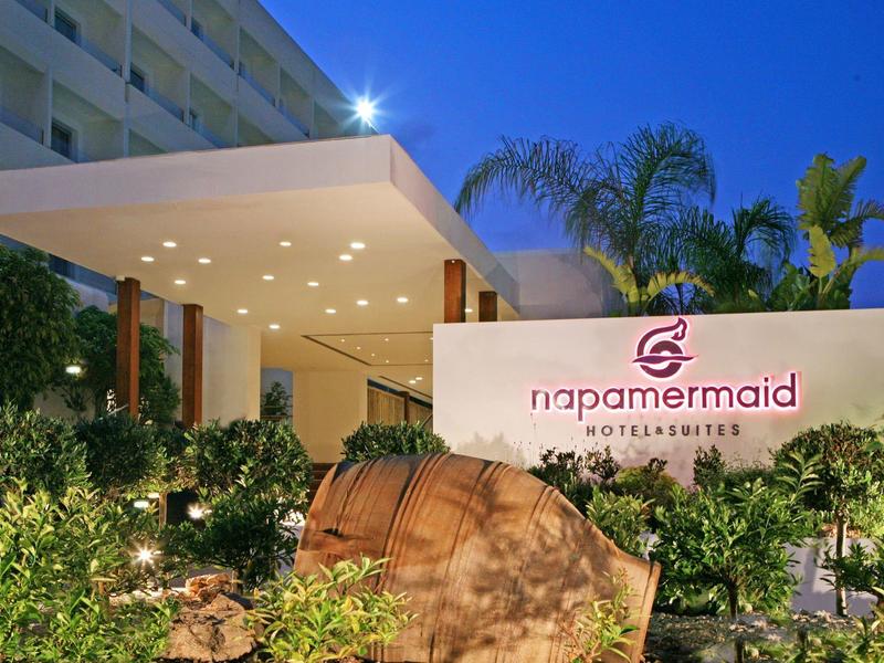 Moderner Eingangsbereich mit beleuchtetem Vordach, Palmen und grünem Pflanzenarrangement bei Abenddämmerung.