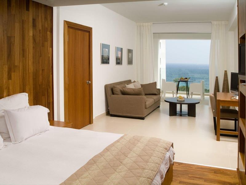 Helles Schlafzimmer mit Holzboden, großem Bett, Sofa, Tisch und Meerblick durch großes Fenster.