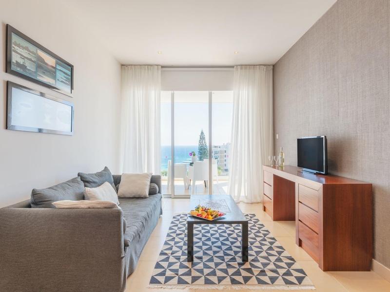 Helles Wohnzimmer mit grauem Sofa, kleinem Tisch, Teppich und Fernsehschrank mit TV vor großem Fenster.