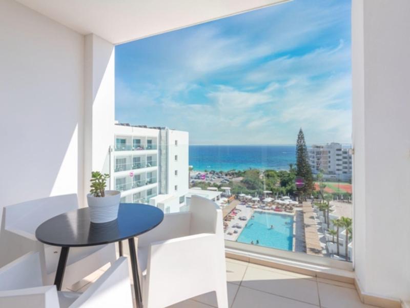 Balkon mit weißem Stuhl, kleinem Tisch und Blumentopf, Blick auf Pool, Gebäude und Meer bei blauem Himmel.