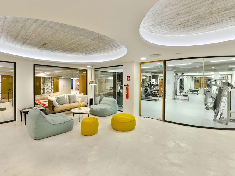 Moderner Lounge-Bereich mit hellen Sofas, runden gelben Hockern und großem Fitnessraum hinter Glaswänden.