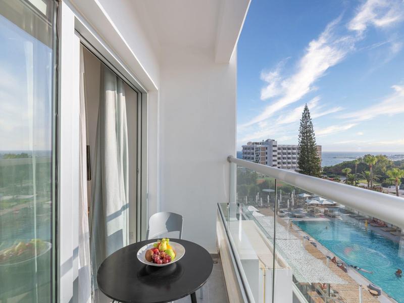 Balkon mit Glastrennwand, schwarzem Tisch mit Obstschale, Blick auf Pool, Bäume und Gebäude am Meer.