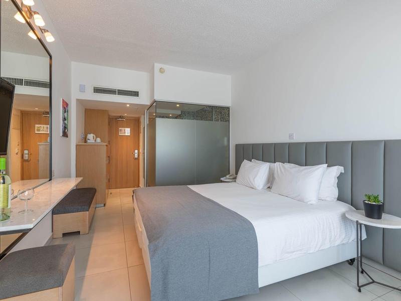 Moderner Hotelzimmer mit großem Bett, grauem Kopfteil, weißen Wänden und minimalistischer Einrichtung.