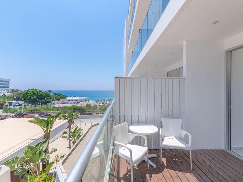 Balkon mit Holzboden, zwei weißen Stühlen und kleinem Tisch, Blick auf Meer und grüne Bäume.