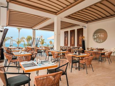 Otevřená restaurace s dřevěnými a ratanovými židlemi, s výhledem na palmy a moře.
