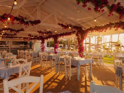 Elegantní restaurace s bílými židlemi a modrými ubrusy, zdobená červenými květinami.