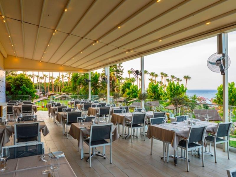Salle à manger extérieure avec tables, chaises et vue sur la mer sous un toit éclairé.