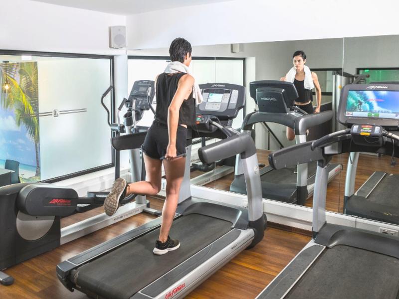 Femme s'entraîne sur tapis roulant dans salle de sport moderne avec miroirs et appareils.