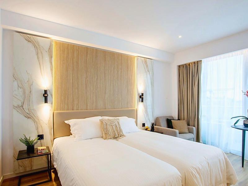 Chambre d'hôtel moderne avec deux lits simples, bois clair, rideaux et canapé près d'une fenêtre.