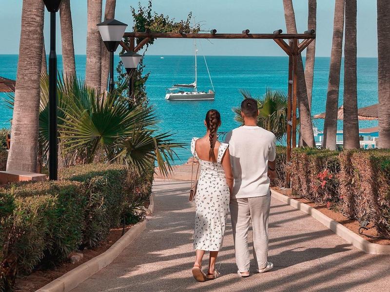 Couple marchant vers la mer turquoise entre des palmiers et des haies bien taillées.