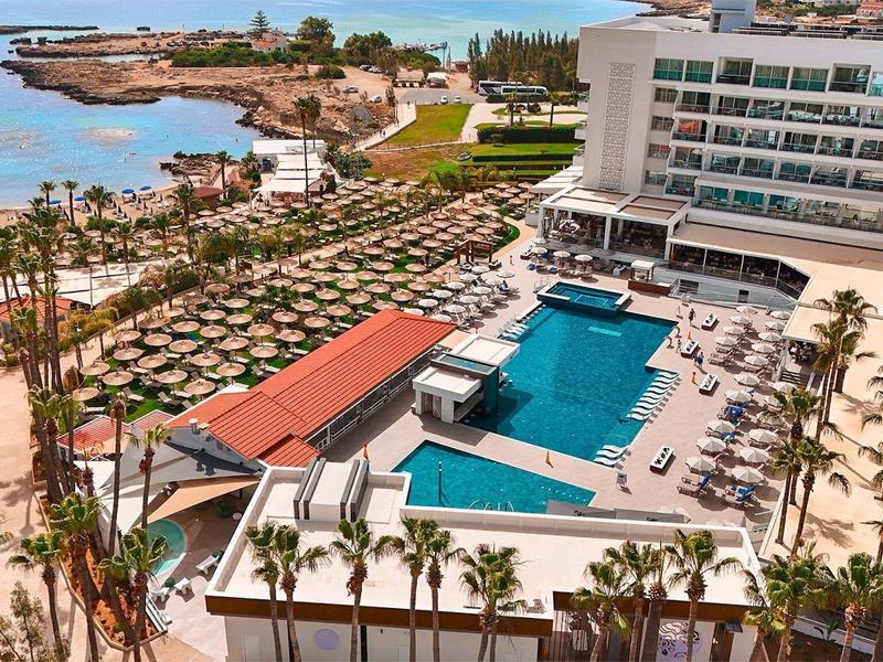 Hôtel de luxe avec piscine, palmiers et vue sur la mer sous un ciel ensoleillé.