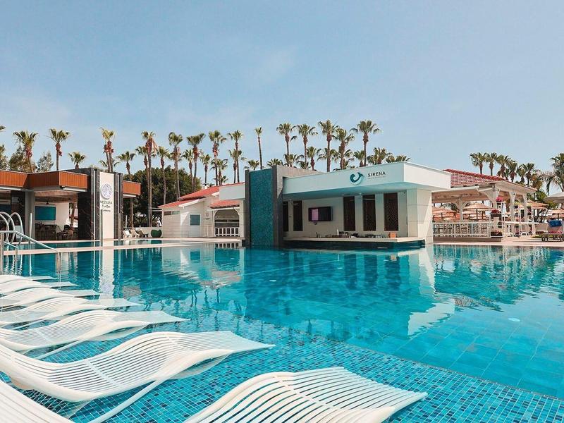 Grande piscine extérieure avec chaises longues blanches et palmiers dans une station balnéaire ensoleillée.