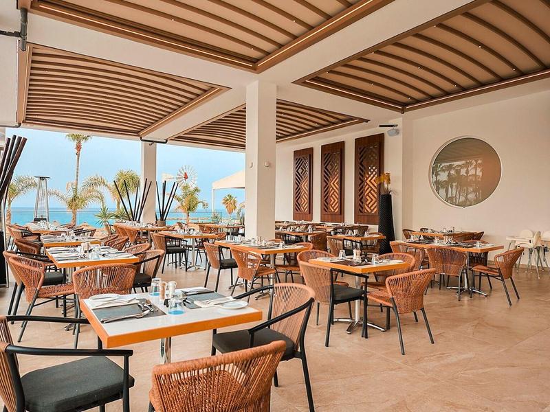 Espace restaurant moderne et ouvert avec des meubles en bois et vue sur les palmiers et la mer.